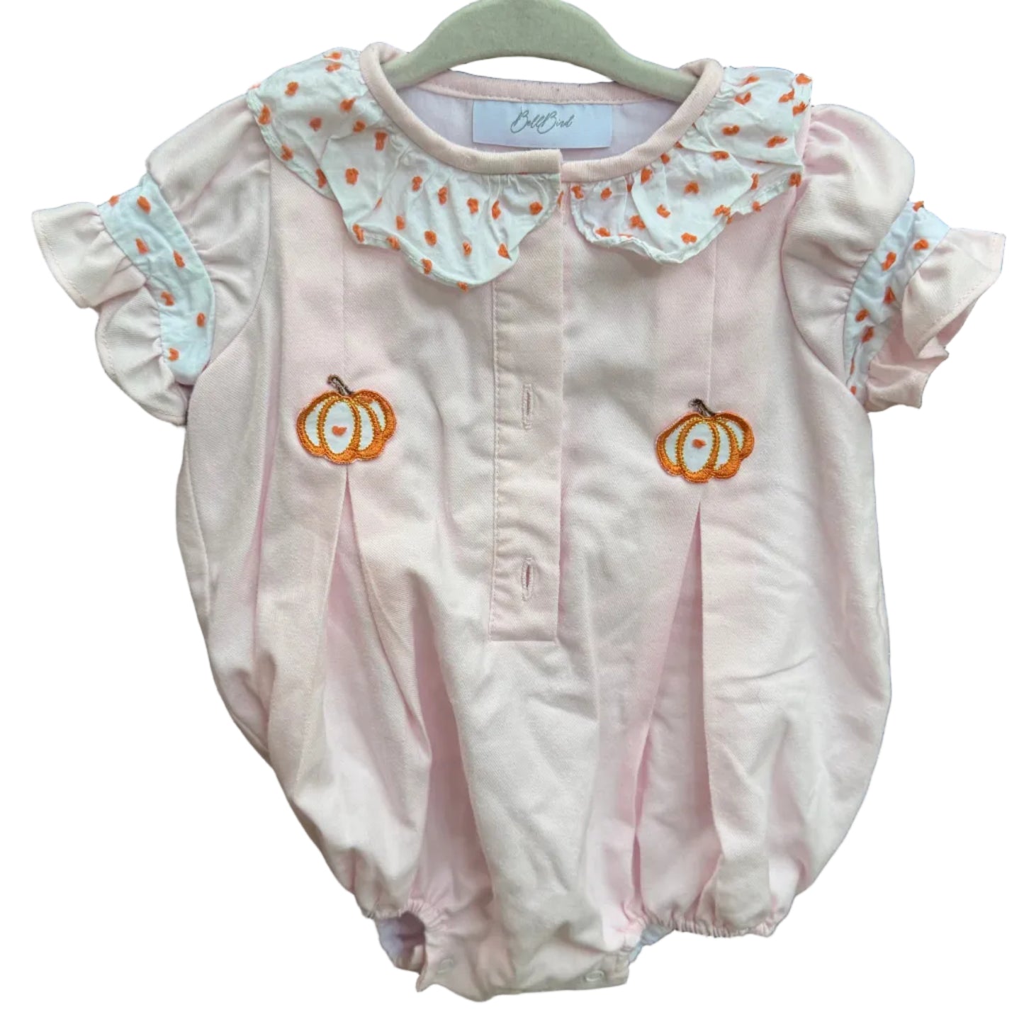 A baby bubble romper in light pink, hanging on a gray hanger against a brick wall. The romper features a white ruffled Peter Pan collar decorated with orange and yellow pumpkin motifs, short flutter sleeves with light blue and white patterned trim, and two embroidered orange pumpkins on the chest.