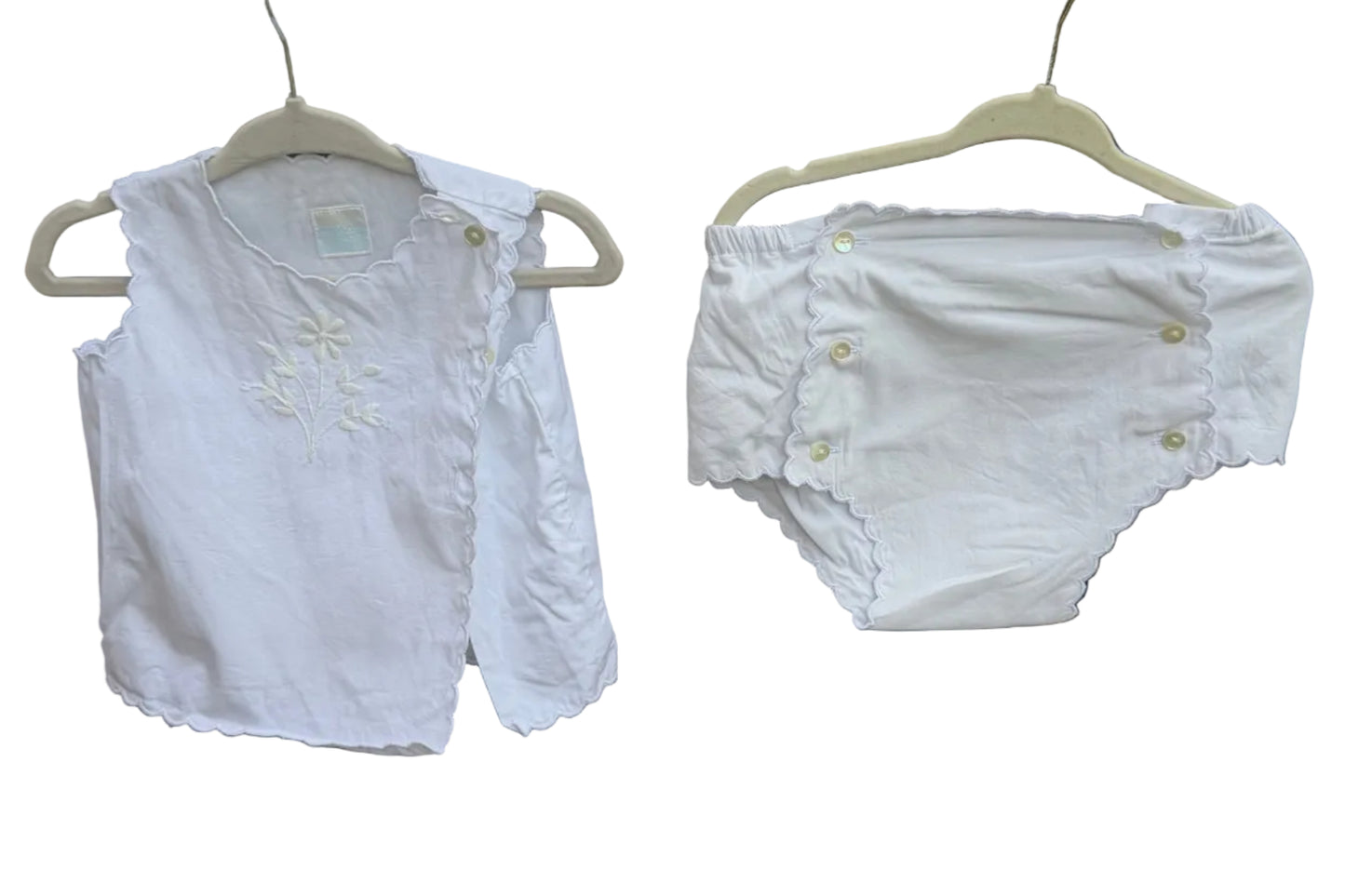 A baby outfit set consisting of a white long-sleeve top with scalloped edges and embroidery, and matching white diaper cover with snap closures, hanging on hangers against a brick wall with trees in the background.