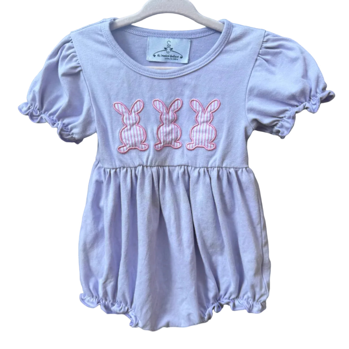 A baby girl's lavender short-sleeve romper with three pink embroidered bunny motifs on the chest, hanging on a white wooden hanger against a brick wall.