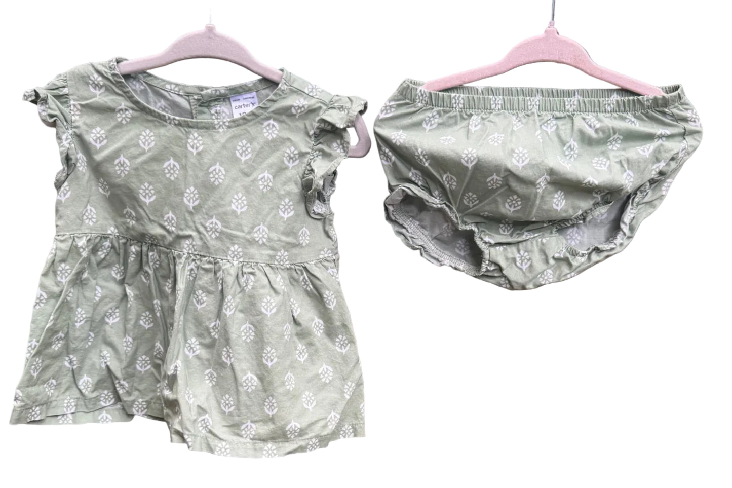 A baby girl's two-piece outfit consisting of a short-sleeve ruffle-sleeve top and matching diaper cover, both in sage green with a white floral pattern, hanging on pink hangers against a red brick wall.