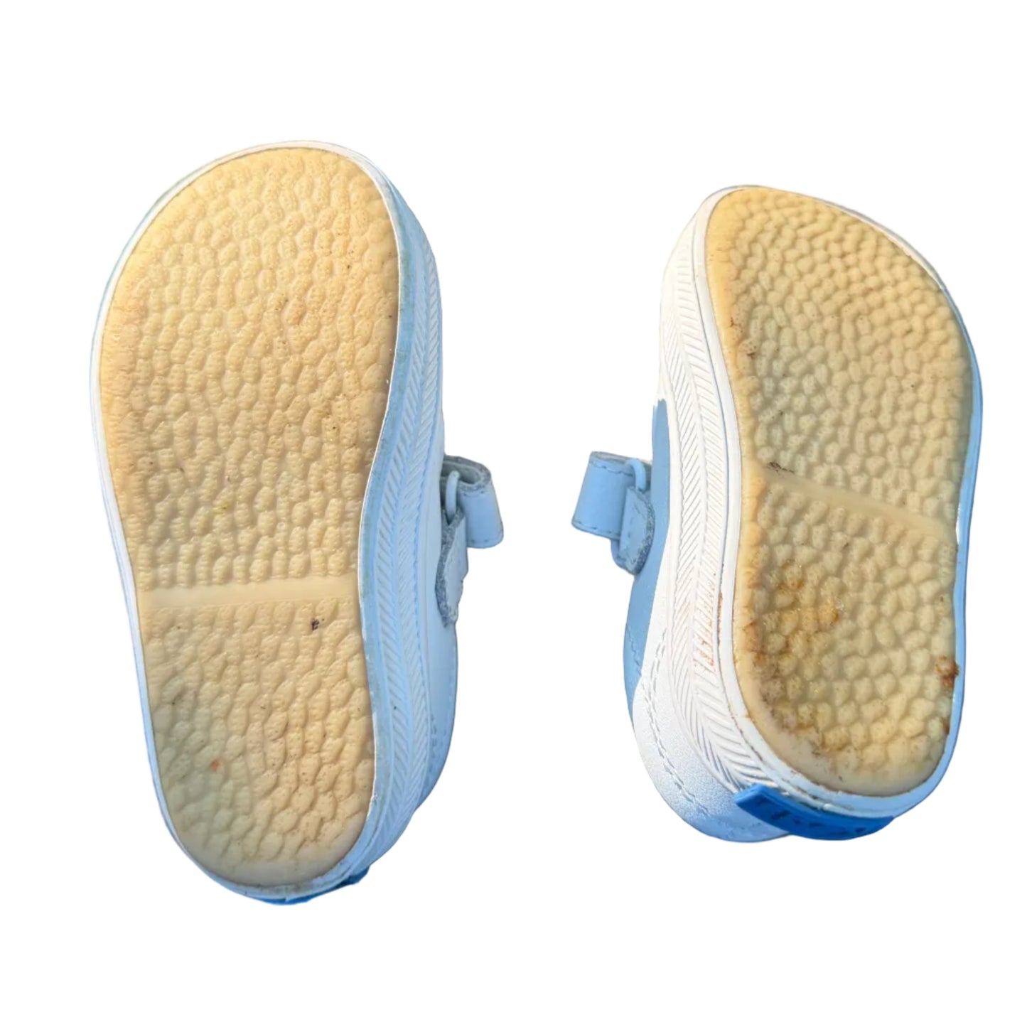 A pair of white children's sneakers shown from the bottom, displaying their heavily worn, yellowed outsole tread. The shoes are resting on weathered brick pavers outdoors, with dirt, grass, and a person's legs visible in the background and foreground.
