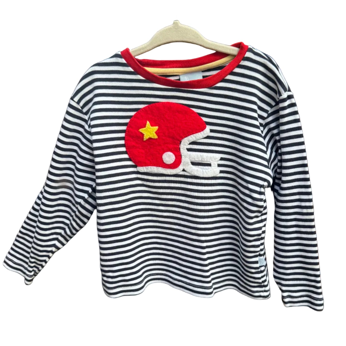 A long-sleeve striped shirt for kids, featuring black and white horizontal stripes, a red crew neck, and a red American football helmet applique with a yellow star on the chest. The shirt is hanging on a wooden hanger against a brick wall background.