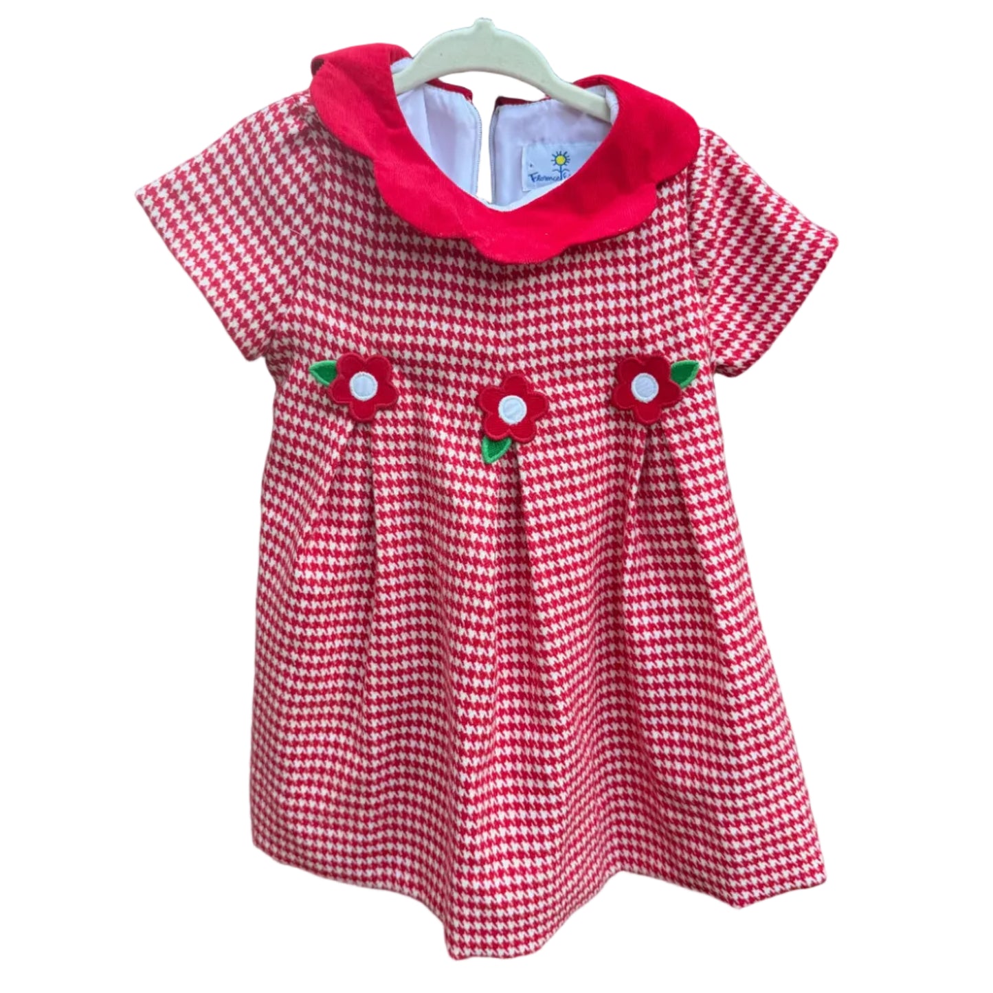 A short-sleeve baby dress with a red and white gingham pattern, a solid red Peter Pan collar, and three strawberry appliques on the front, hanging on a white plastic hanger against a brick wall.