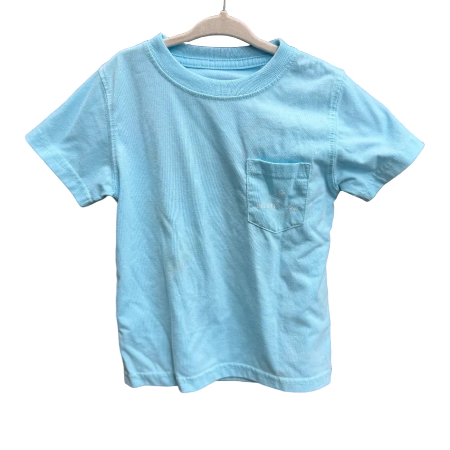 A light blue short-sleeve pocket t-shirt hanging on a wooden hanger against a red-brown brick wall.