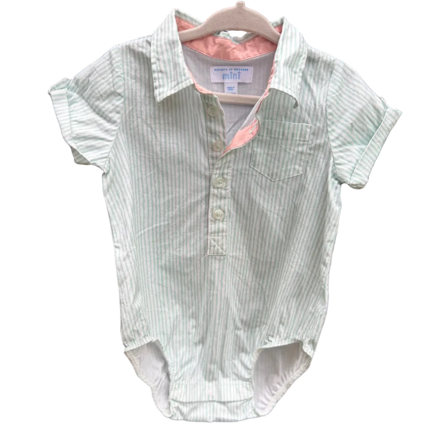 A baby short-sleeve button-front romper with vertical light green and white stripes, hanging on a wooden hanger against a red-brown brick wall.