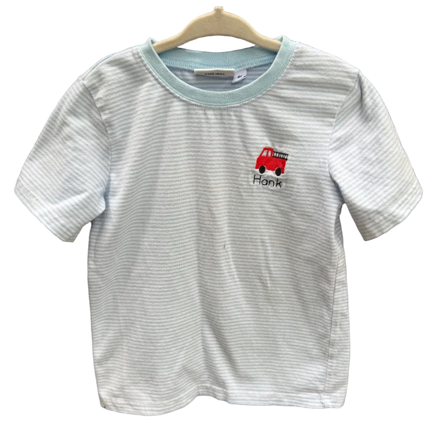 A light blue and white striped short-sleeve t-shirt with a light blue collar, featuring a small red fire truck graphic and the name 'Frank' printed on the chest, hanging on a wooden hanger against a brick wall.