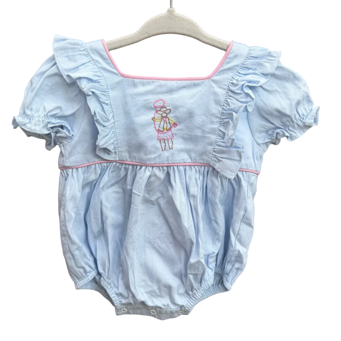 A baby girl's light blue bubble romper with ruffled sleeves and a small embroidered girl in a pink dress on the chest, hanging on a white wooden hanger against a red-brown brick wall.