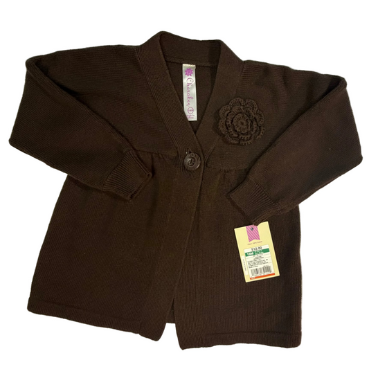 A dark brown cardigan sweater for kids, laid flat on a white sheet. The cardigan features a single button closure, a decorative fabric flower applique on the left chest, and a yellow retail hangtag attached to the sleeve.