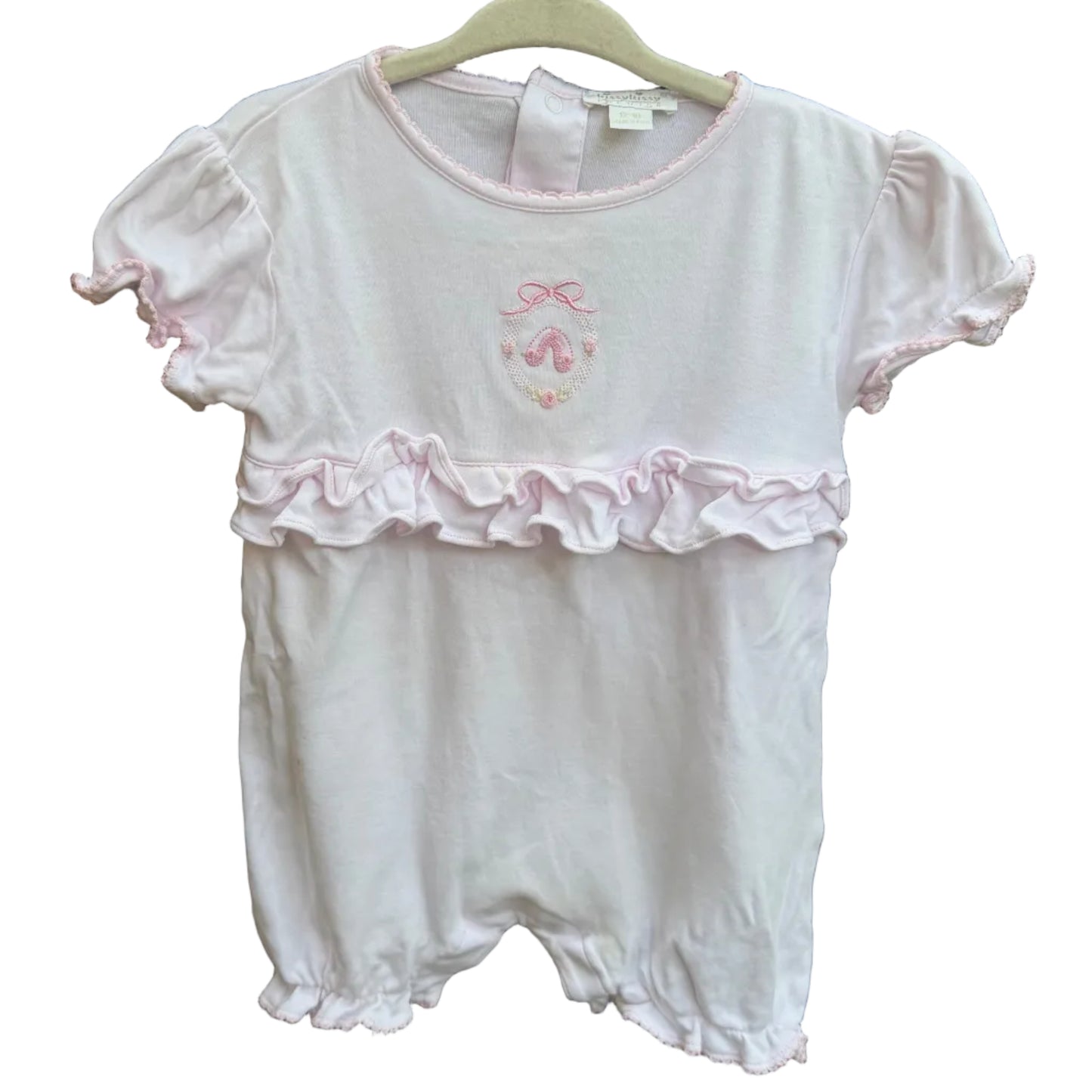 A baby girl's short-sleeve romper in white with a light pink ruffle trim and a small pink bow graphic on the chest, hanging on a white plastic hanger against a brick wall.