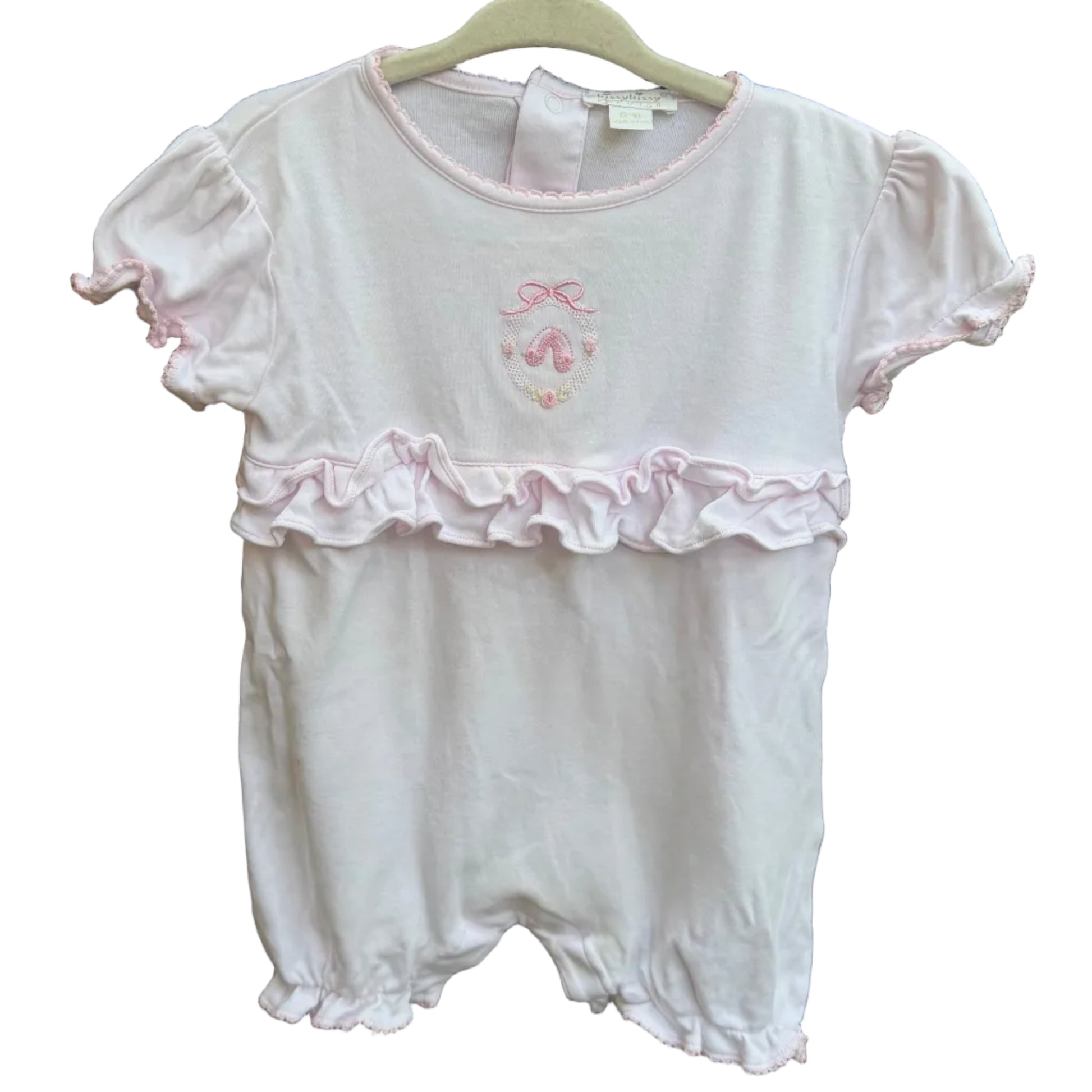 A baby girl's short-sleeve romper in white with a light pink ruffle trim and a small pink bow graphic on the chest, hanging on a white plastic hanger against a brick wall.