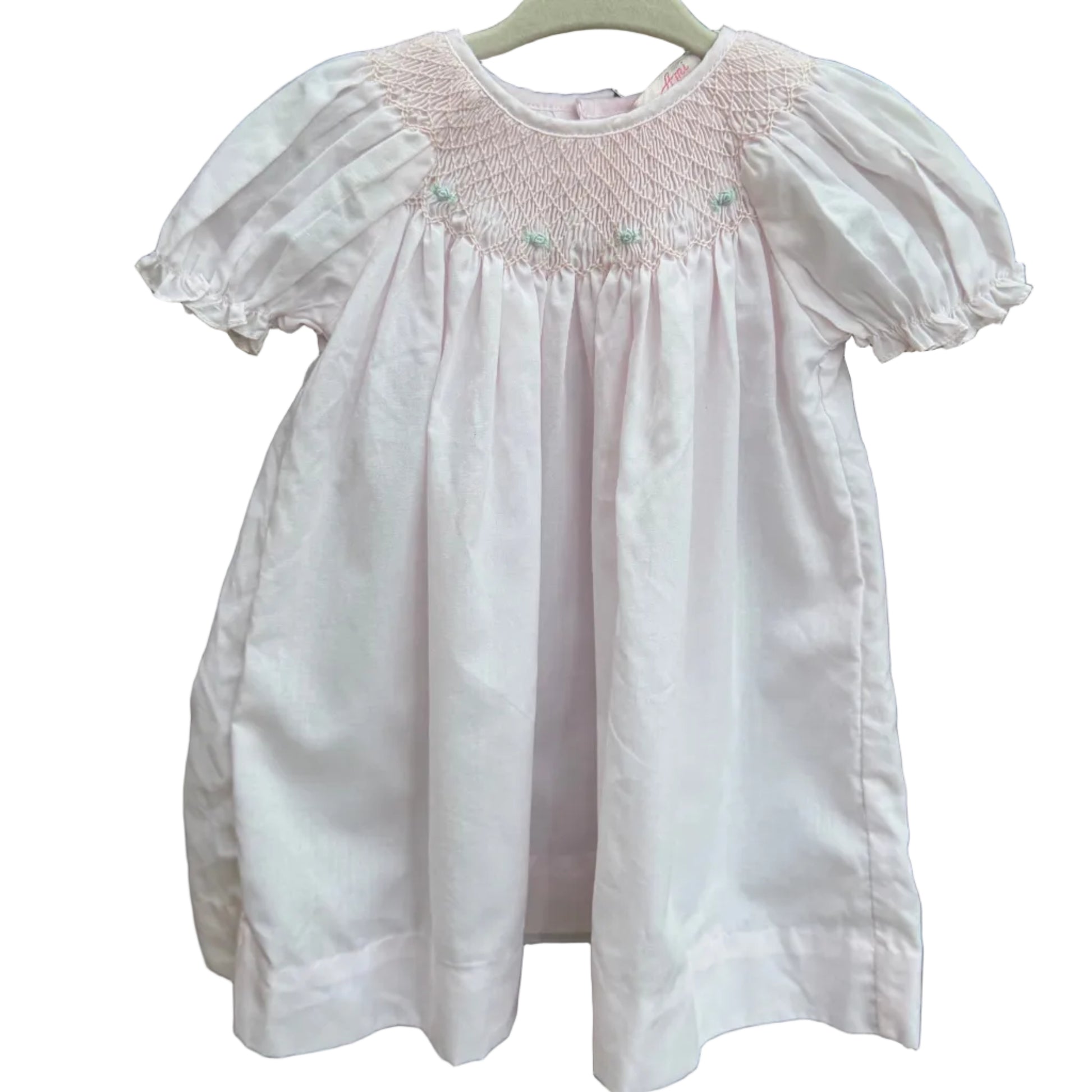 A light pink baby smocked dress with short puff sleeves hanging on a white plastic hanger against a brown brick wall with green foliage in the background.