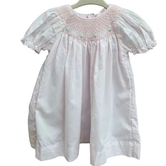 A light pink baby smocked dress with short puff sleeves hanging on a white plastic hanger against a brown brick wall with green foliage in the background.