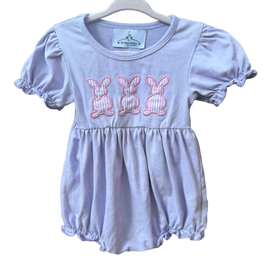 A baby girl's lavender short-sleeve romper with three pink embroidered bunny motifs on the chest, hanging on a white wooden hanger against a brick wall.