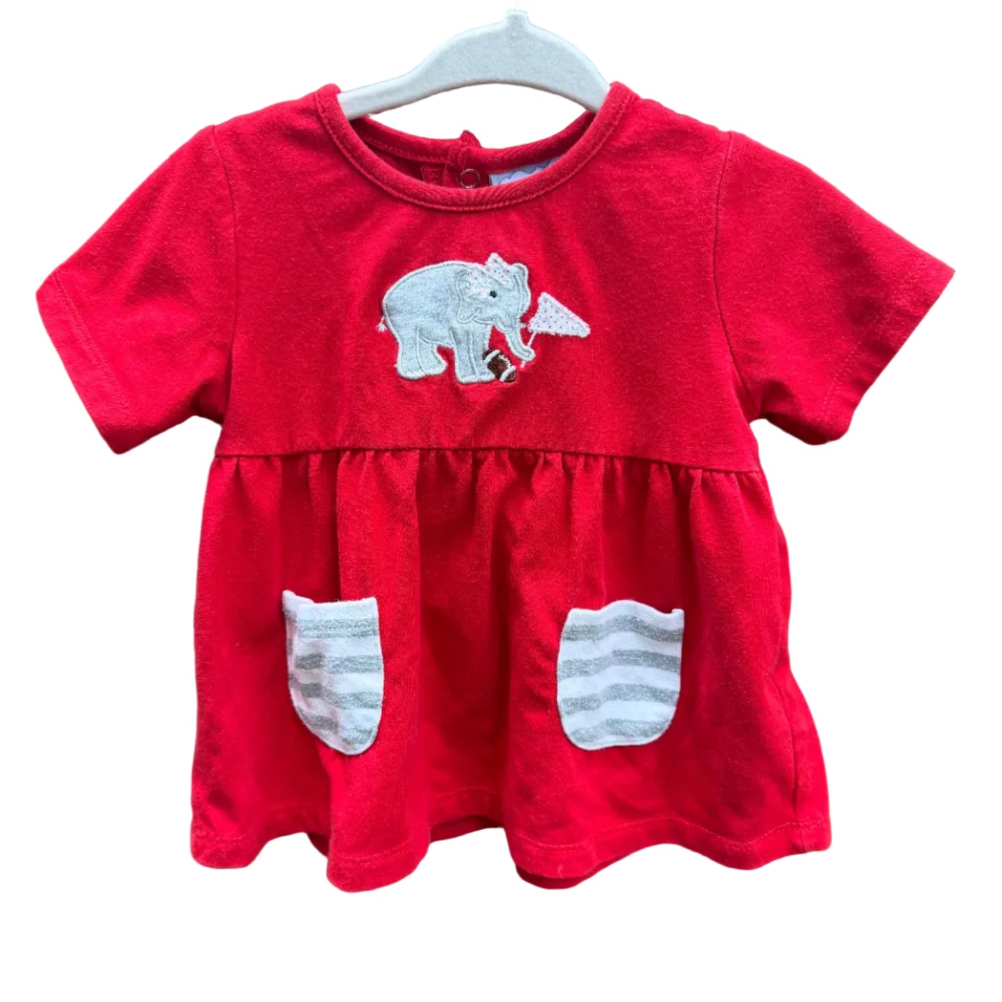 A red short-sleeve baby dress with a white polar bear applique on the chest and two white striped patch pockets, hanging on a white hanger against a brown brick wall.