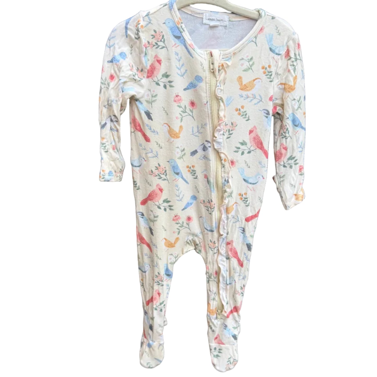 A baby footed one-piece sleeper with a colorful bird and floral print on a cream background, hanging on a wooden hanger against a brown brick wall.