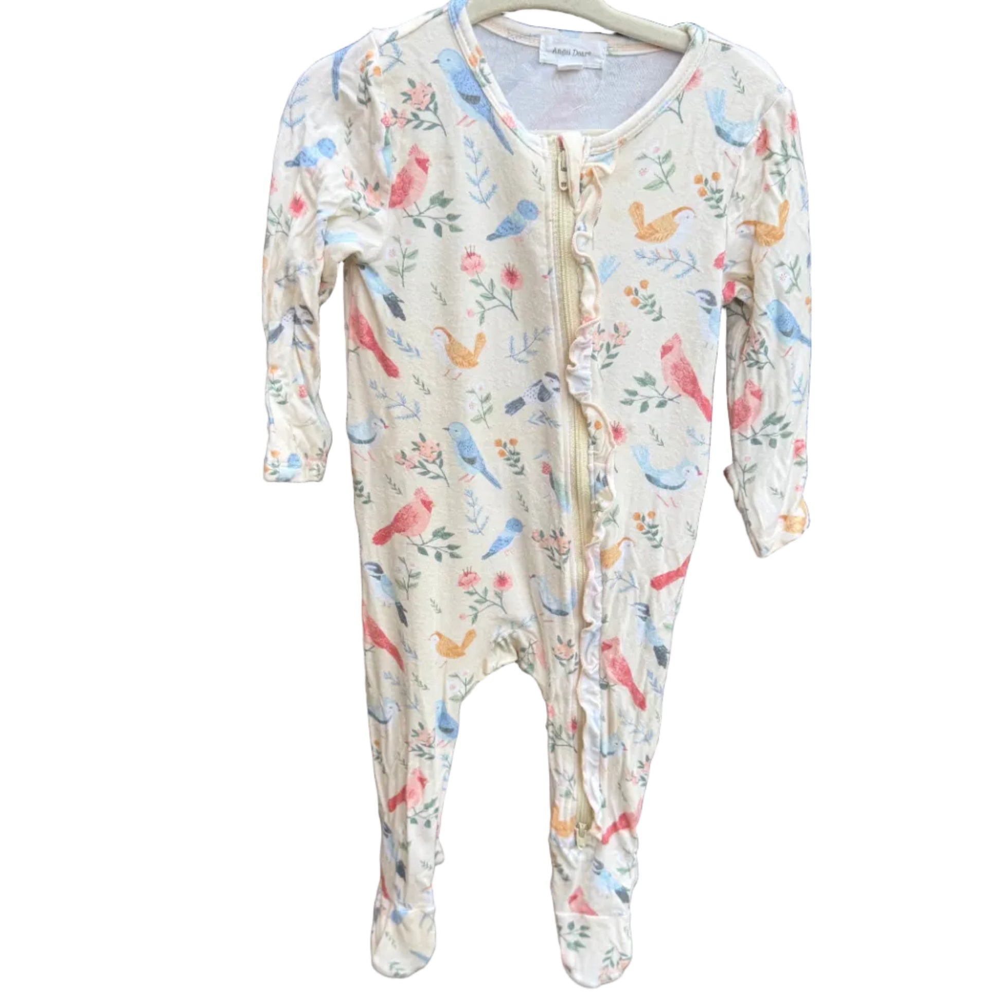 A baby footed one-piece sleeper with a colorful bird and floral print on a cream background, hanging on a wooden hanger against a brown brick wall.