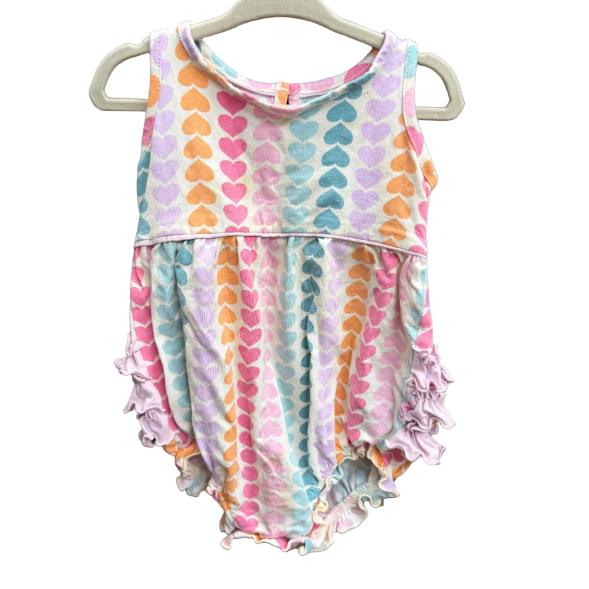 A sleeveless baby romper with a pastel heart pattern and ruffled trim, hanging on a white plastic hanger against a brown brick wall.