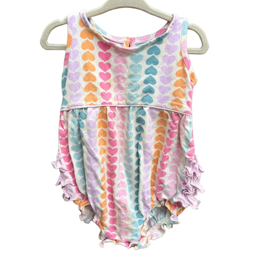 A sleeveless baby romper with a pastel heart pattern and ruffled trim, hanging on a white plastic hanger against a brown brick wall.