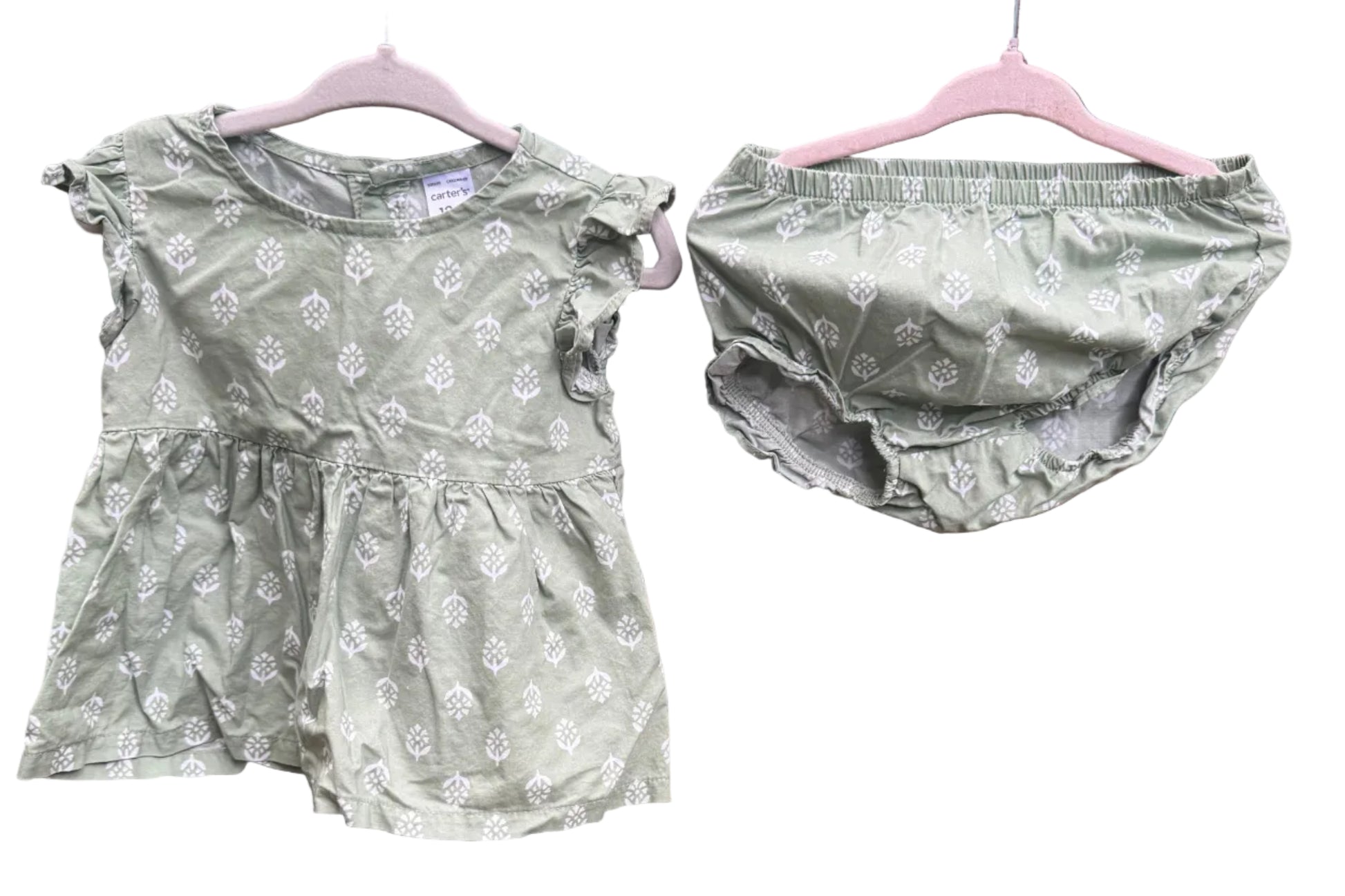 A baby girl's two-piece outfit consisting of a short-sleeve ruffle-sleeve top and matching diaper cover, both in sage green with a white floral pattern, hanging on pink hangers against a red brick wall.