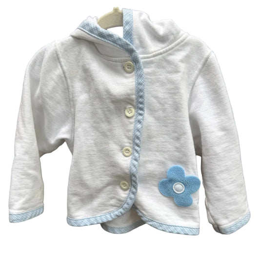 A white long-sleeve baby cardigan with light blue trim, snap buttons, and a blue flower applique, laid flat on a brown brick background.