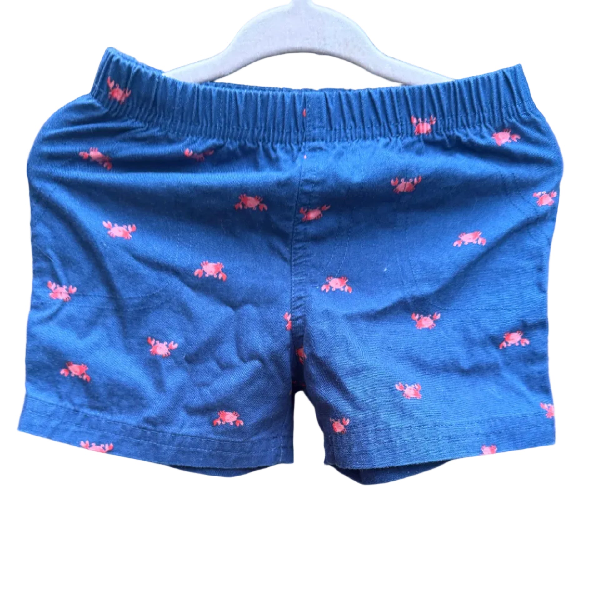 Blue children's shorts with a pink lobster print, hanging on a white plastic hanger against a brick wall background.