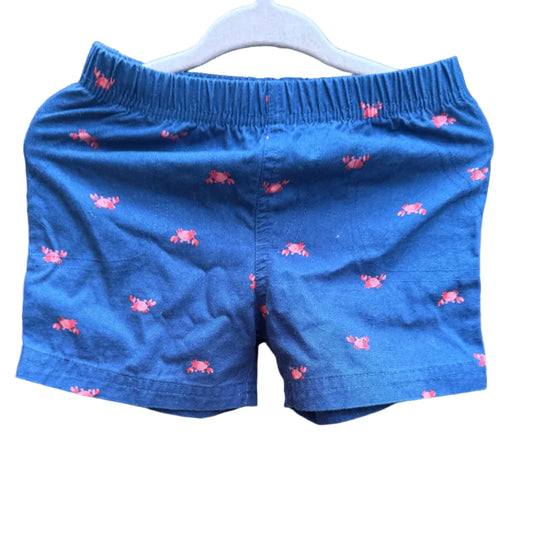 Blue children's shorts with a pink lobster print, hanging on a white plastic hanger against a brick wall background.