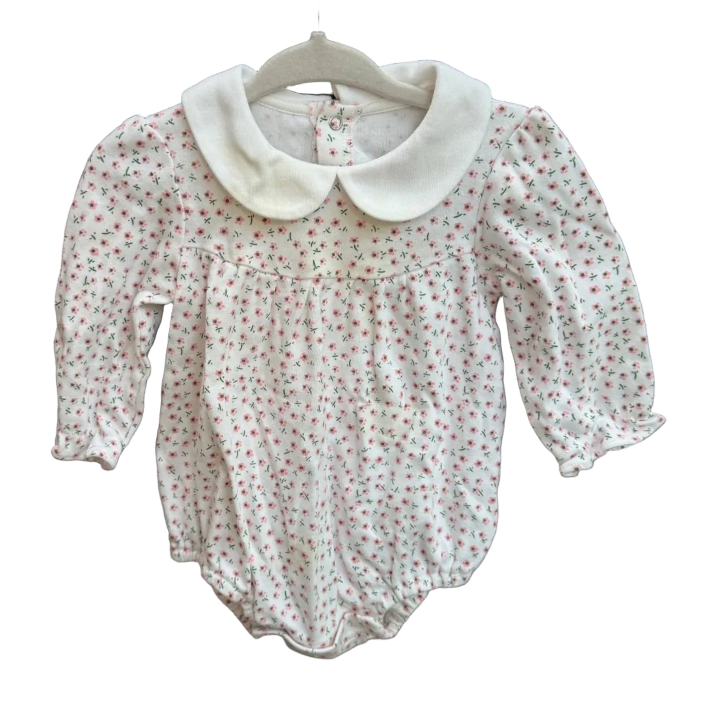 A baby girl's long-sleeve romper in white fabric with a tiny red and green floral print, featuring a large white Peter Pan collar, hanging on a white hanger against a brick wall.