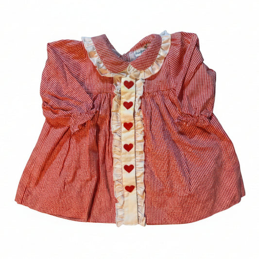 A red and white gingham baby dress with a cream ruffled collar, cuffs, and center placket decorated with red hearts, laid flat on a beige fabric-covered surface.