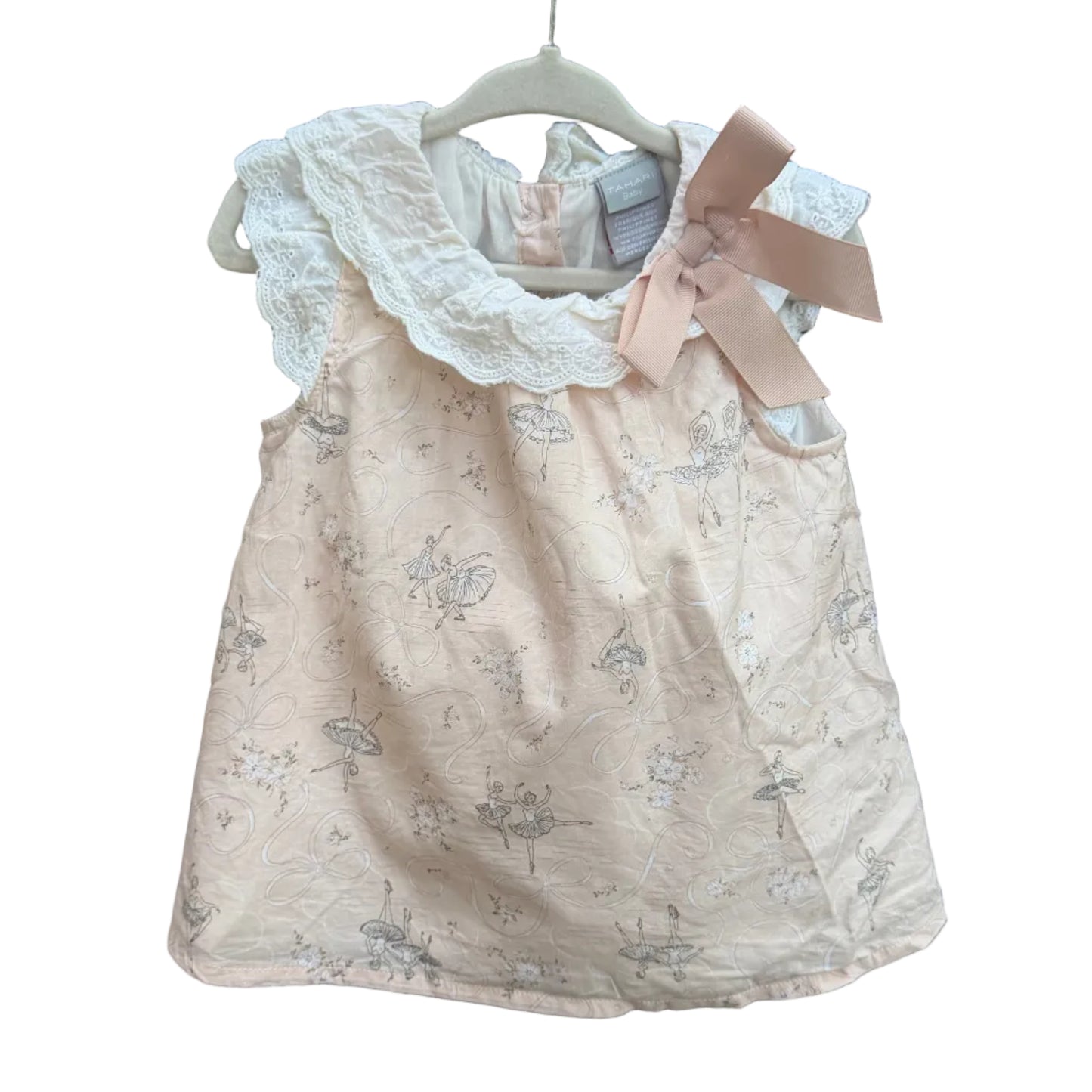 A light pink sleeveless baby dress with a delicate gray floral print, featuring a white lace collar and a large dusty pink bow on the shoulder, hanging on a brick wall.