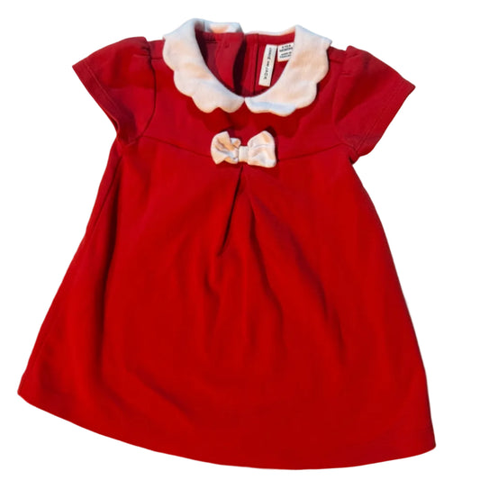 A red baby girl's dress with a white ruffled Peter Pan collar and a matching bow on the chest, laid flat on a light-colored fabric surface.