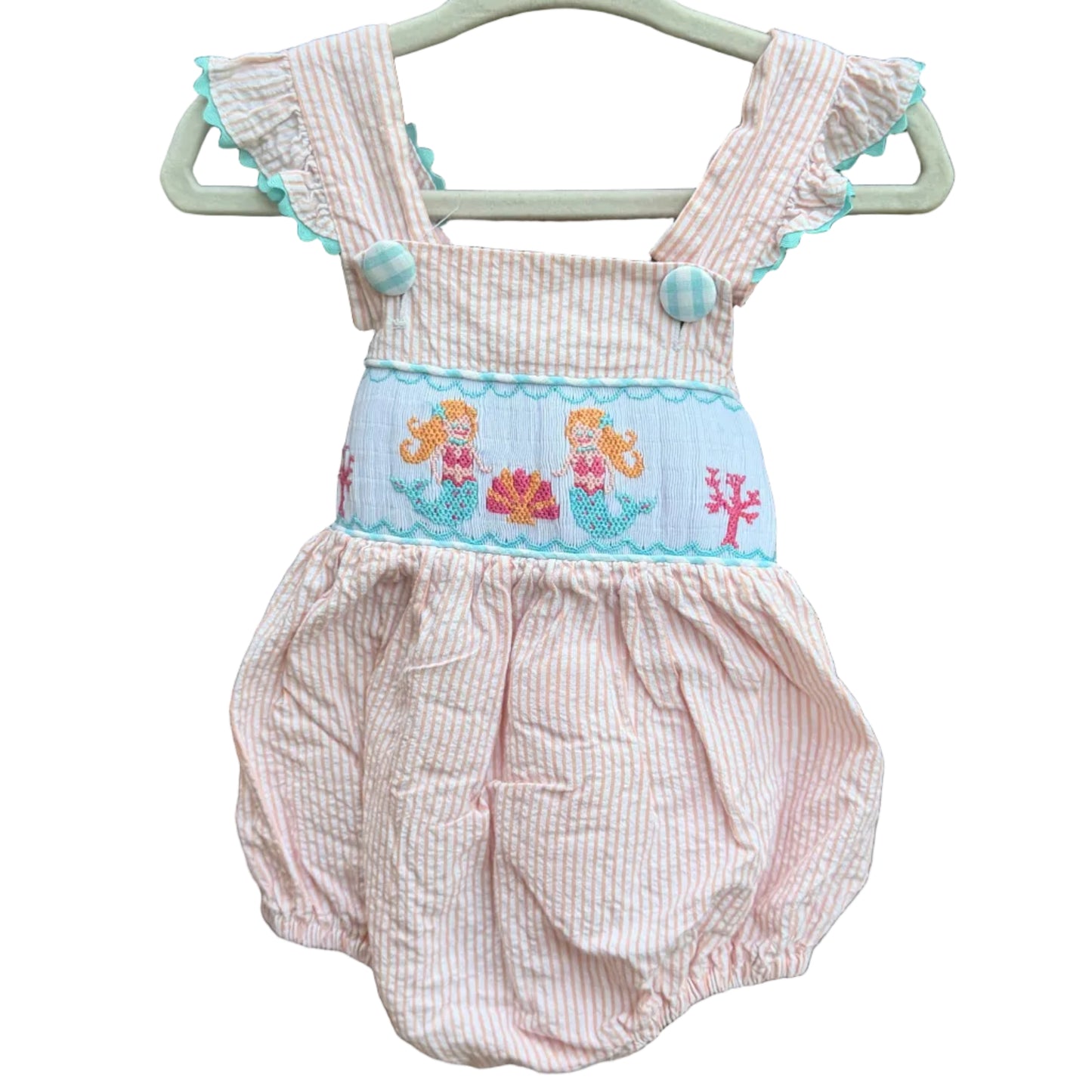 A orange and white seersucker baby romper with a light blue smocked mermaid panel, hanging on a white plastic hanger against a brick wall.