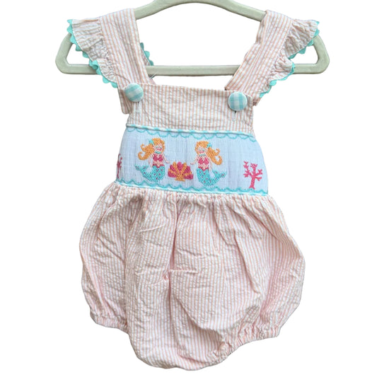 A orange and white seersucker baby romper with a light blue smocked mermaid panel, hanging on a white plastic hanger against a brick wall.