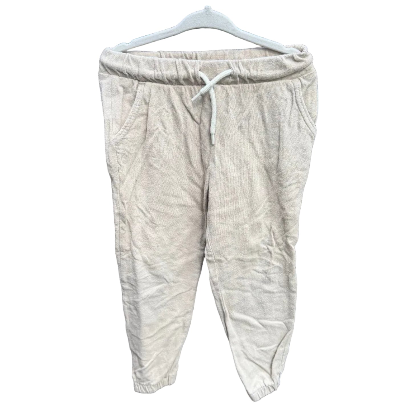 A pair of light beige linen jogger pants hanging on a white plastic hanger against a red-brown brick wall with green foliage visible above it.