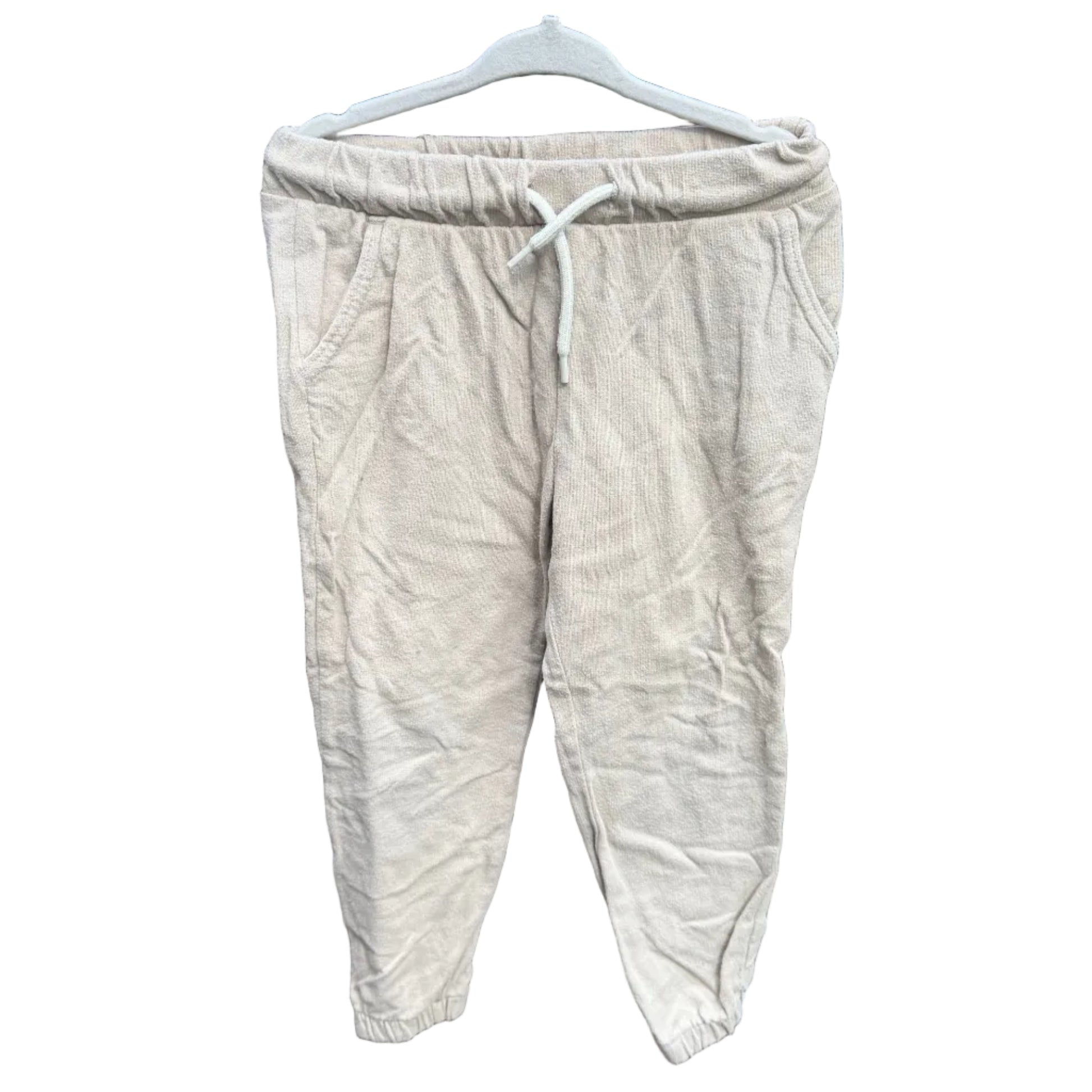 A pair of light beige linen jogger pants hanging on a white plastic hanger against a red-brown brick wall with green foliage visible above it.