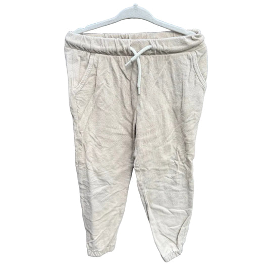 A pair of light beige linen jogger pants hanging on a white plastic hanger against a red-brown brick wall with green foliage visible above it.