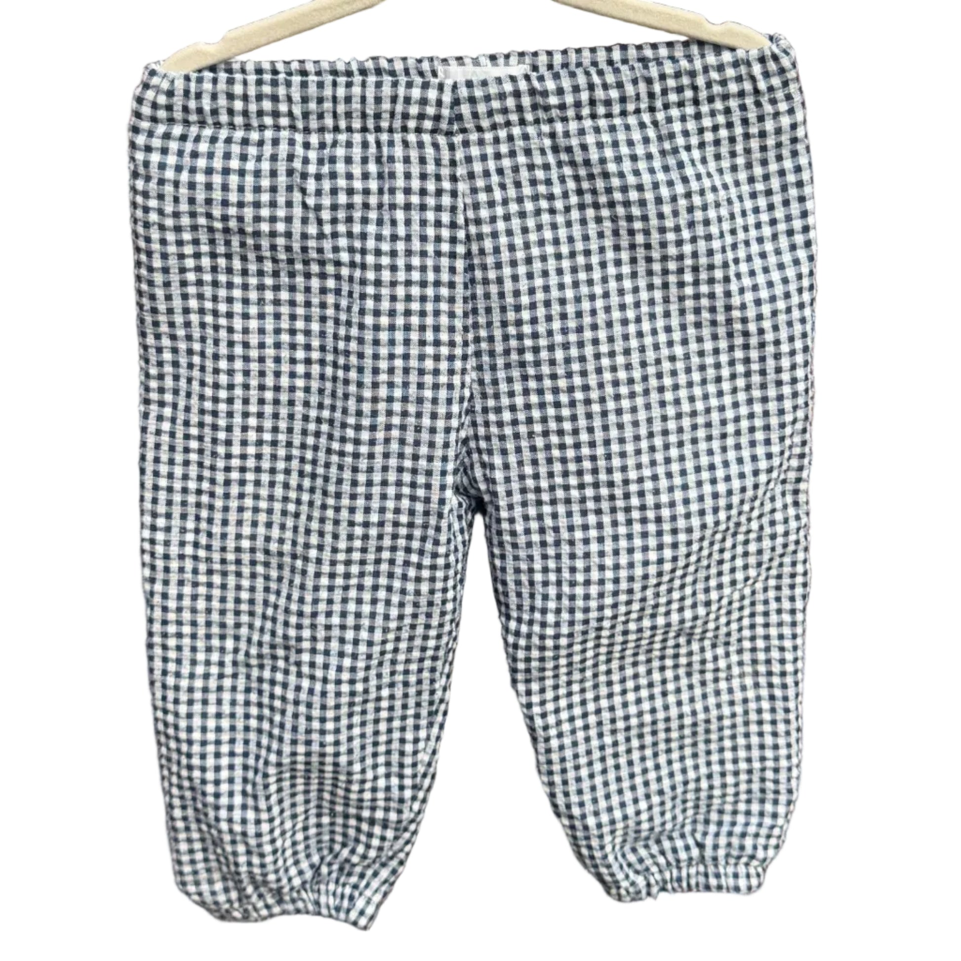 A pair of children's gingham pants in a black and white checkered pattern, hanging on a wooden hanger against a brick wall with green trees in the background.
