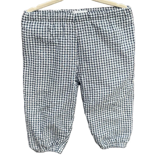 A pair of children's gingham pants in a black and white checkered pattern, hanging on a wooden hanger against a brick wall with green trees in the background.
