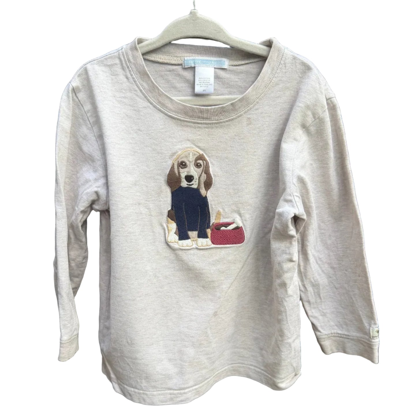 A light beige long-sleeve t-shirt with a graphic of a beagle dog wearing a black sweater and holding a red bag, hanging on a white hanger against a brick wall background.