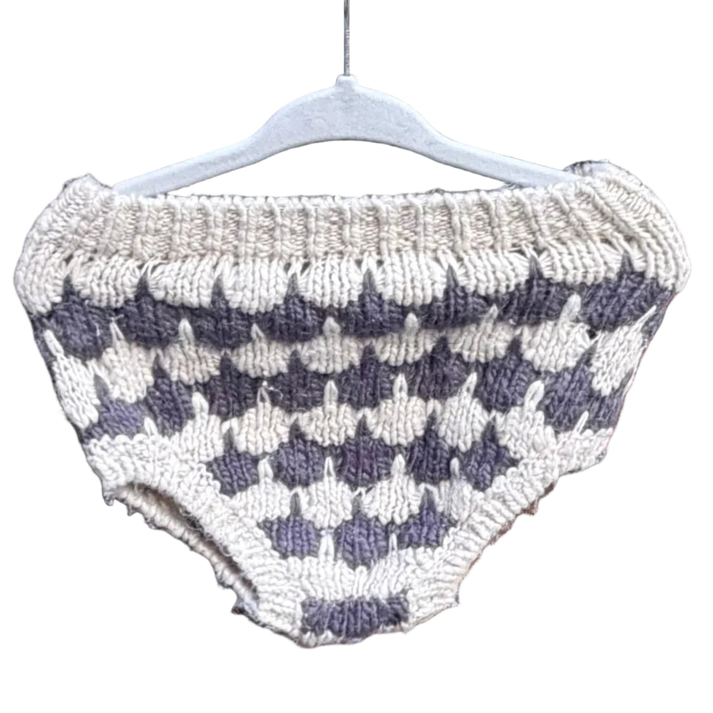 A hand-knit baby diaper cover or shorts in a cream and dark gray checkerboard pattern, hanging on a white plastic hanger against a red-brown brick wall with trees in the background.