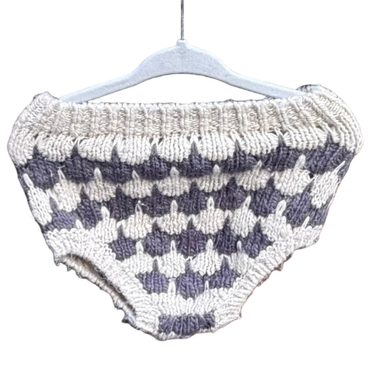 A hand-knit baby diaper cover or shorts in a cream and dark gray checkerboard pattern, hanging on a white plastic hanger against a red-brown brick wall with trees in the background.