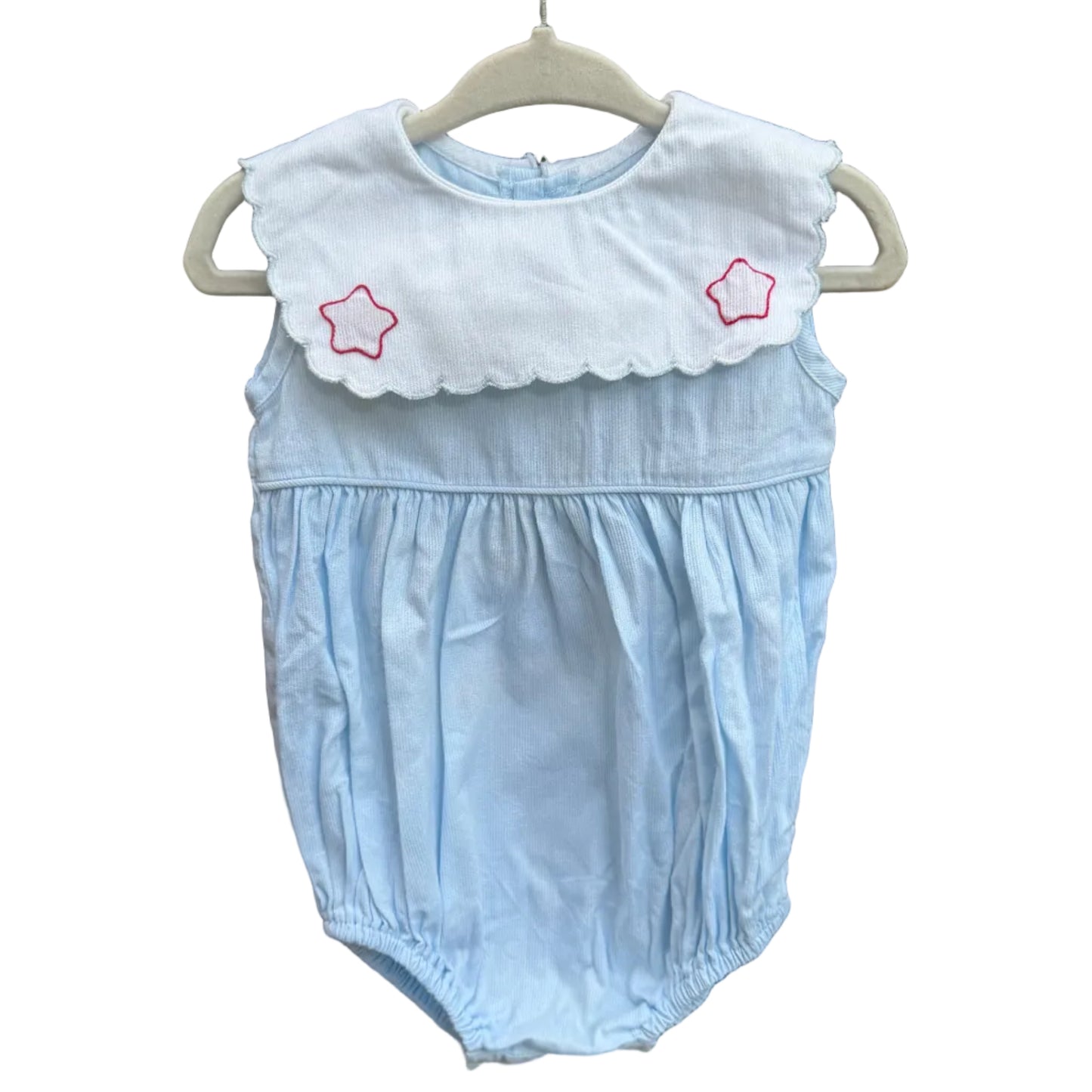 A light blue baby romper with a white ruffled collar featuring two embroidered red stars, hanging on a white plastic hanger against a red-brown brick wall.