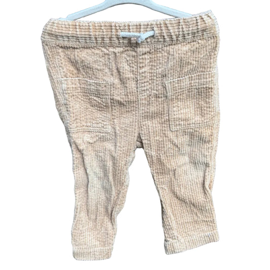 A pair of light beige textured knit pants hanging on a white plastic hanger against a red-brown brick wall with green foliage visible above.