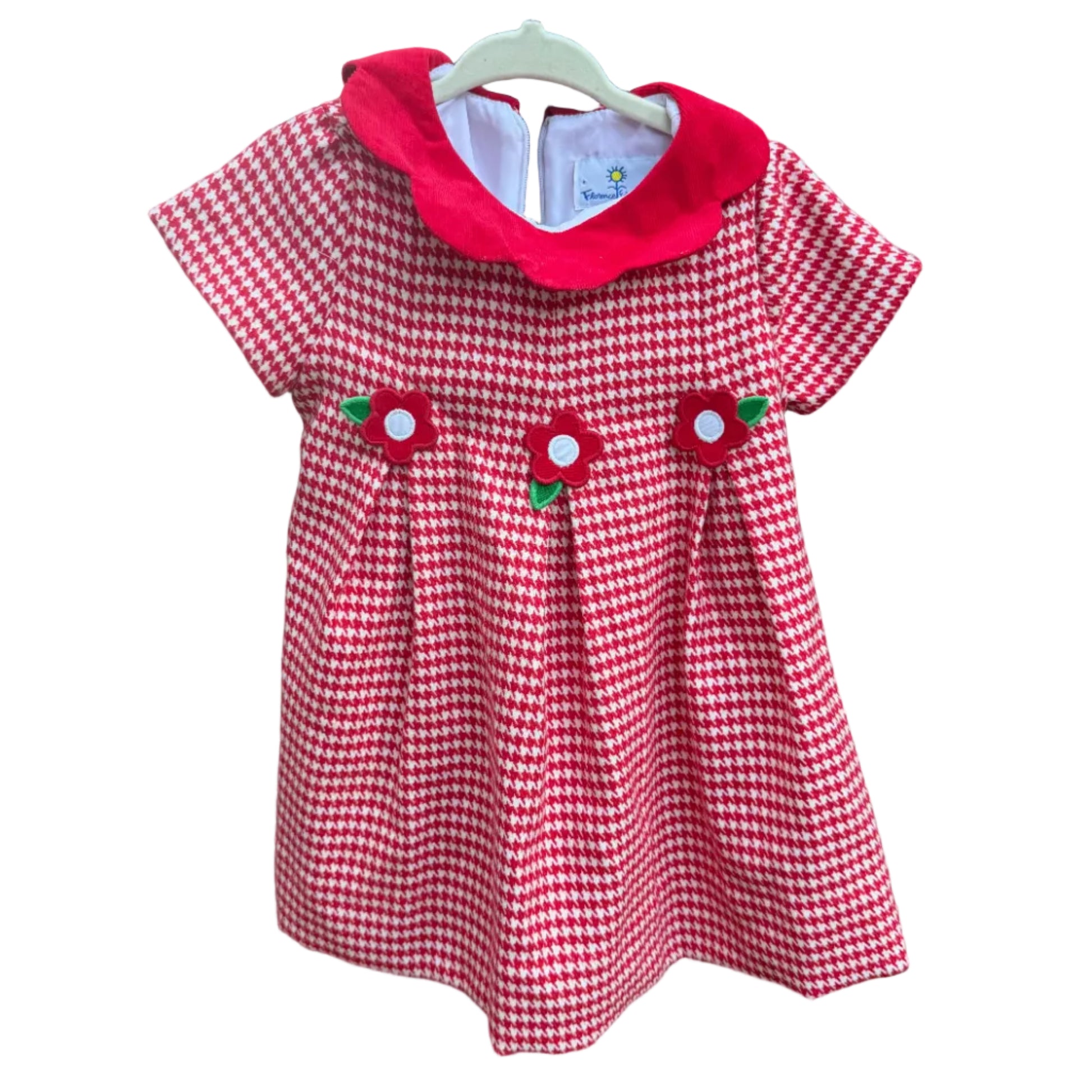 A short-sleeve baby dress with a red and white gingham pattern, a solid red Peter Pan collar, and three strawberry appliques on the front, hanging on a white plastic hanger against a brick wall.