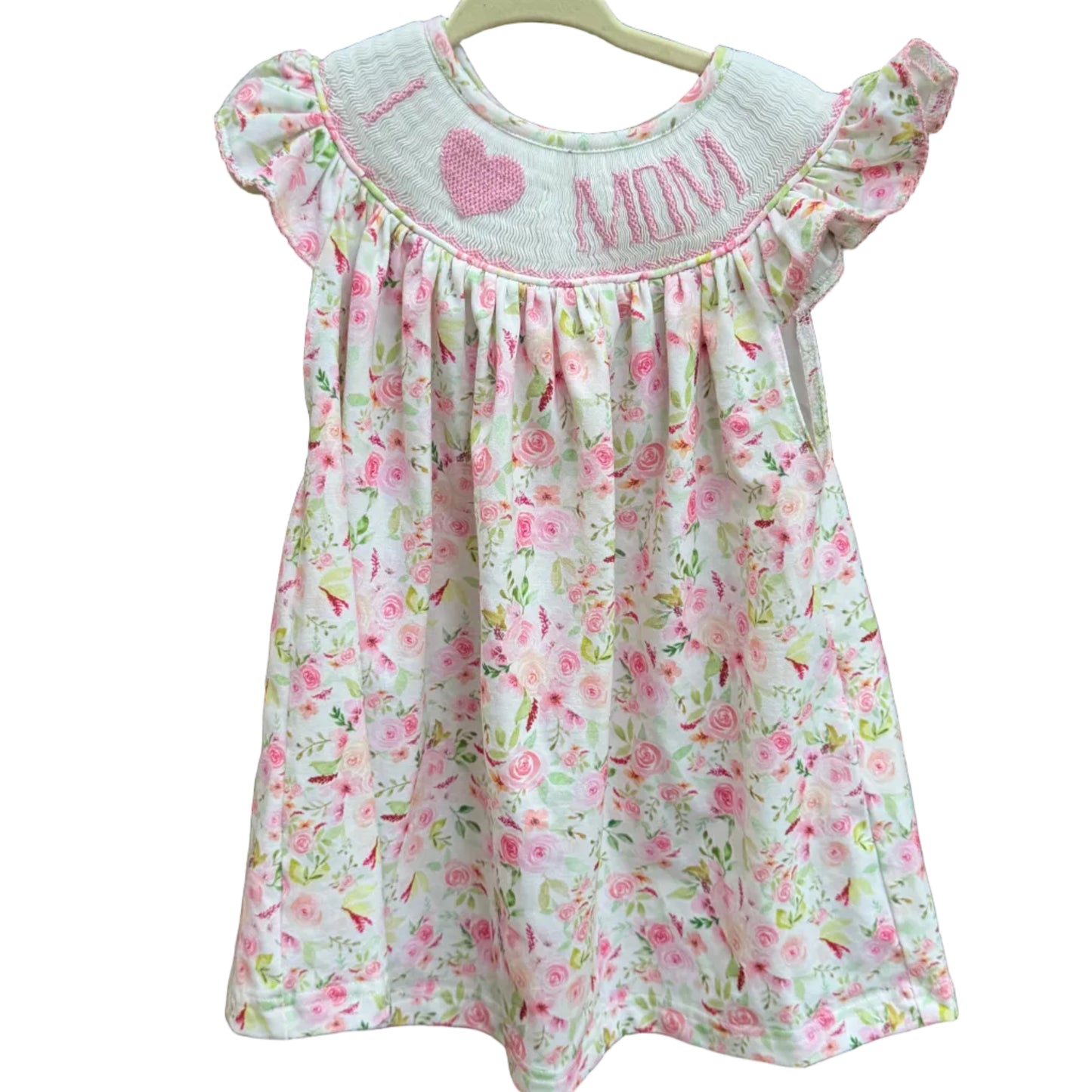 A toddler girl's floral dress with ruffle sleeves hangs on a wooden hanger against a brick wall. The dress has a white base with a pink and green floral pattern, featuring 'I <heart> ANNA' embroidered in pink on the chest.