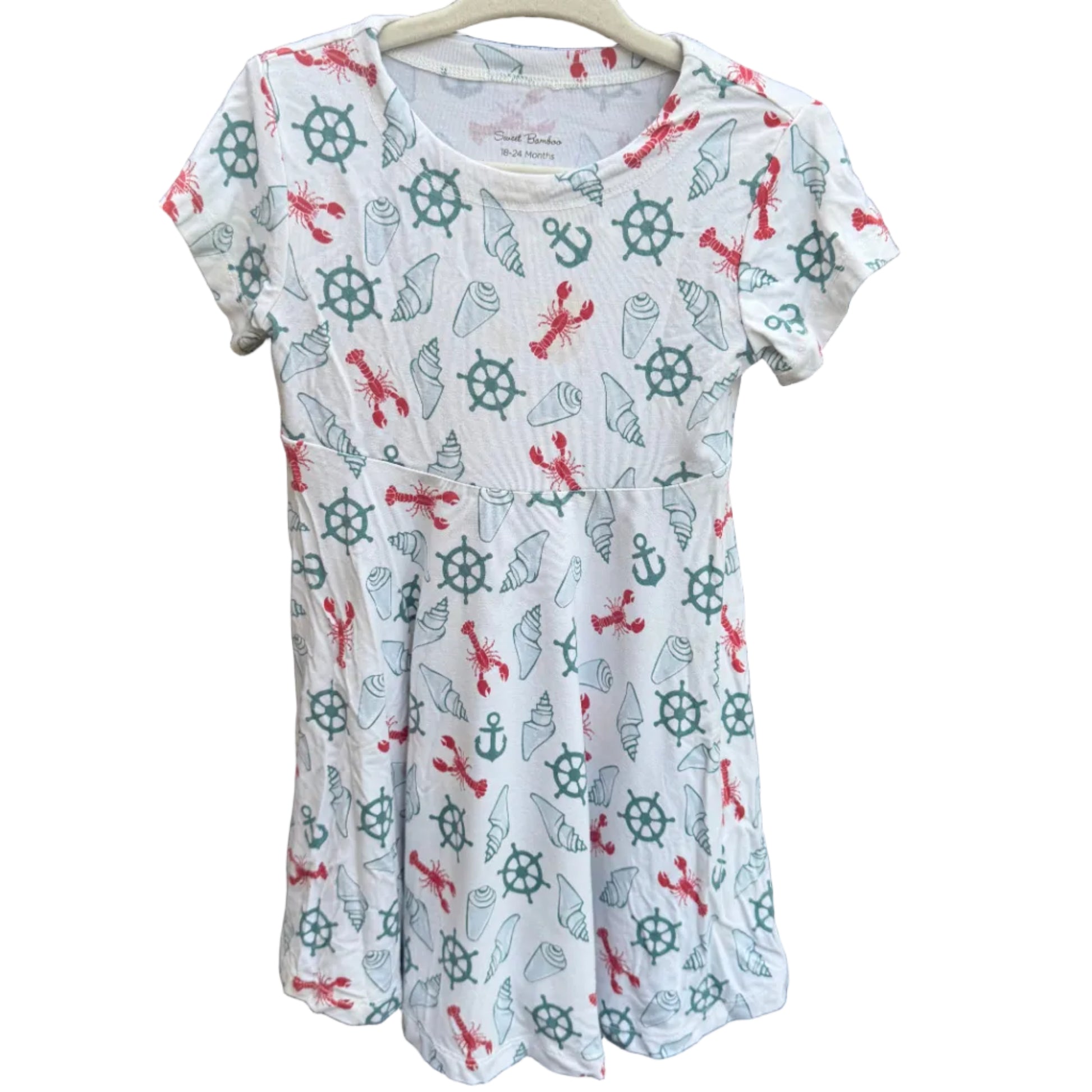 A white short-sleeve t-shirt with a nautical-themed print of anchors, ship wheels, and red crosses, hanging on a wooden hanger against a brown brick wall.