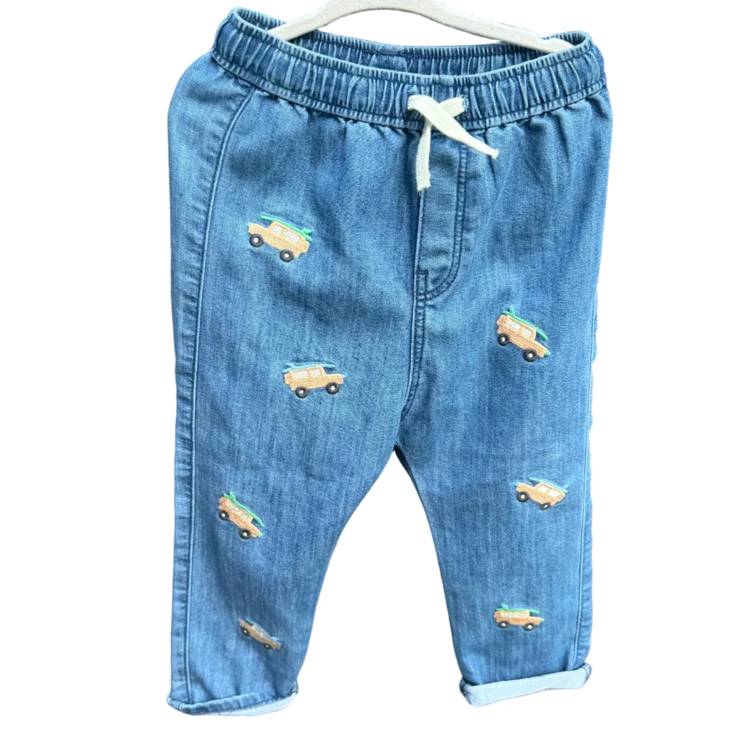 Light blue denim baby pants with embroidered toy cars, hanging on a white hanger against a brick wall background.