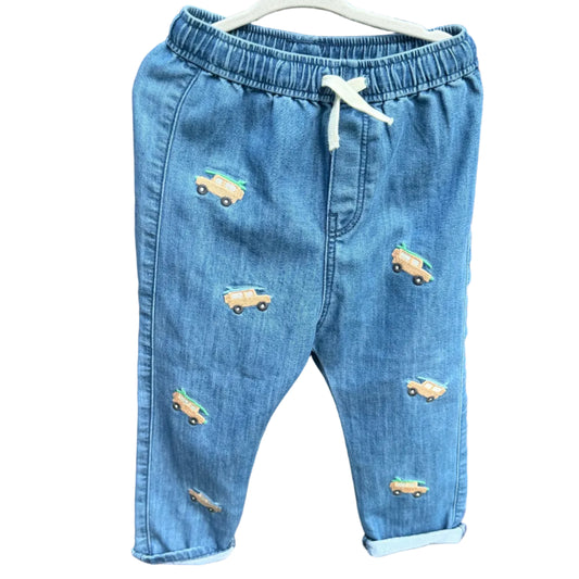 Light blue denim baby pants with embroidered toy cars, hanging on a white hanger against a brick wall background.