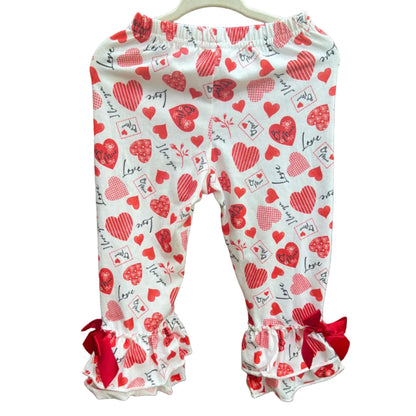 White toddler leggings with a red heart and script pattern, featuring ruffled cuffs with red bows, hanging on a white plastic hanger against a brown brick wall.