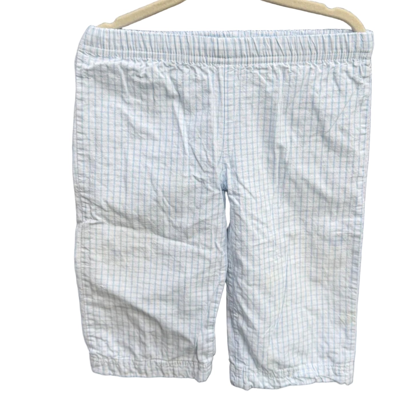 Light blue and white checkered shorts hanging on a beige hanger against a red-brown brick wall with green foliage in the background.