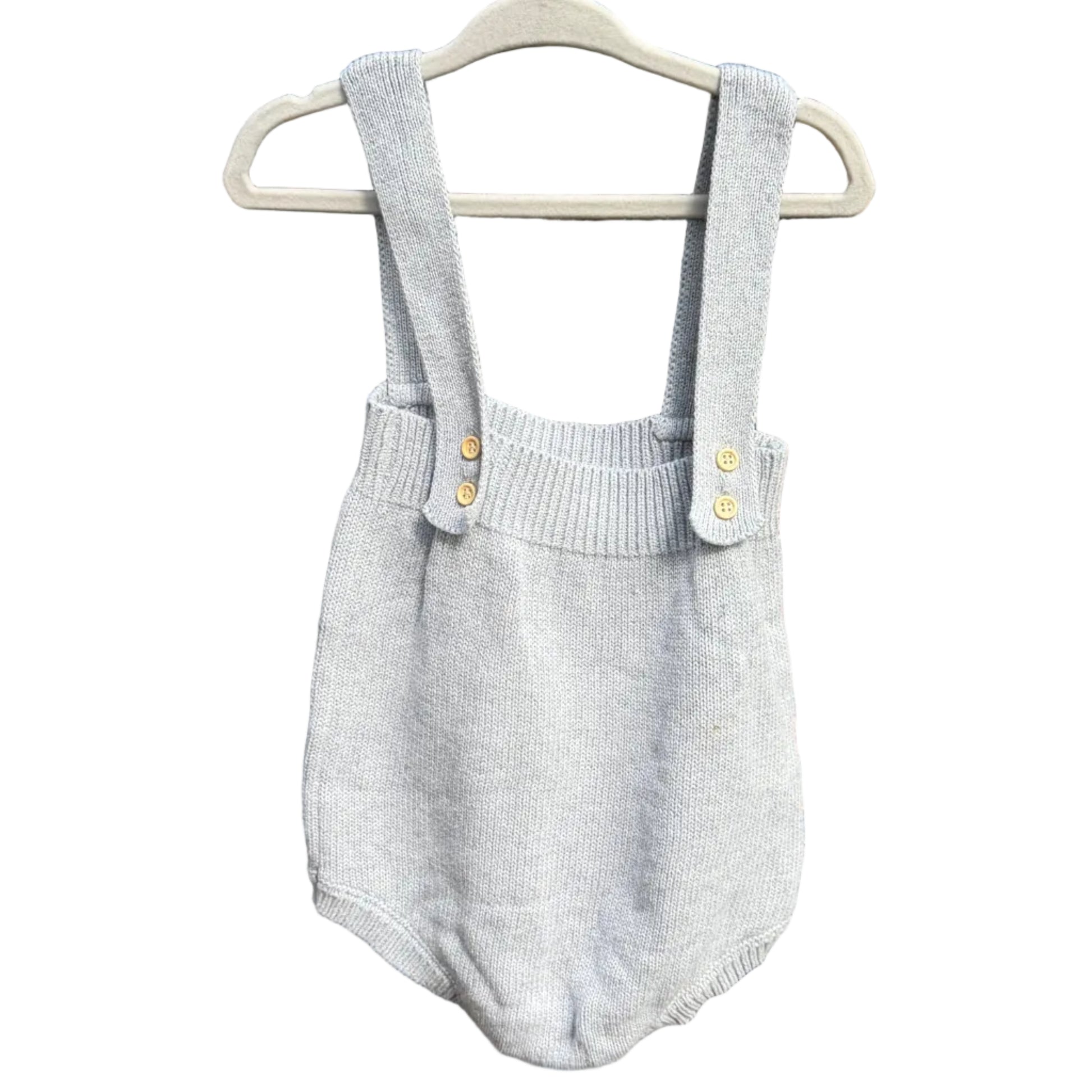 A light gray knit baby romper with shoulder straps and small gold buttons, hanging on a white plastic hanger against a rustic red-brown brick wall.